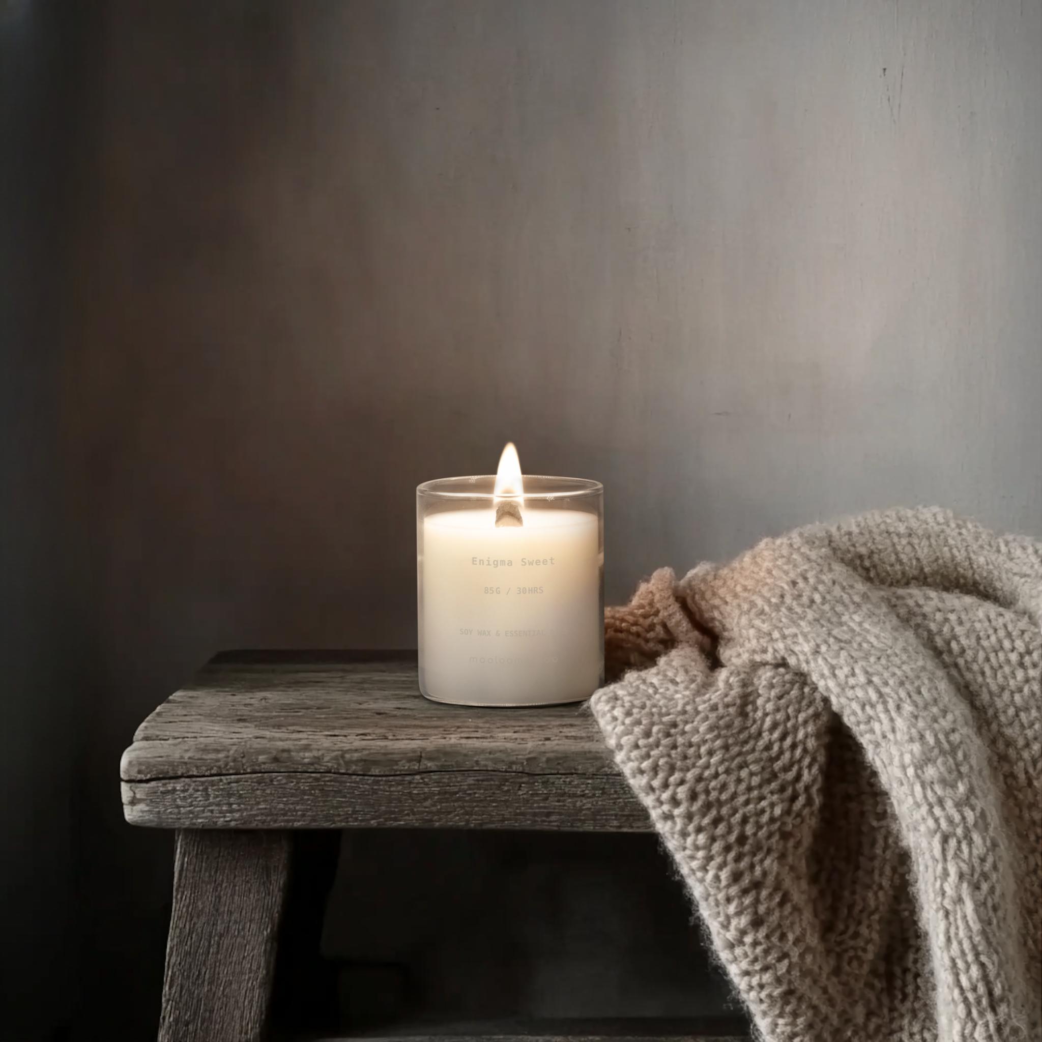 Enigma Sweet candle glowing on wooden bench with cozy blanket – soy wax scented candle